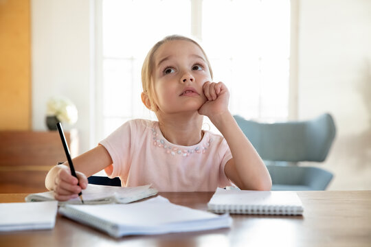 Dreamy small Caucasian girl child sit at desk feel unmotivated doing home task assignment alone. Thoughtful little kid look in distance dreaming thinking, lazy preparing homework. Learning concept.
