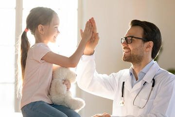 Happy young Caucasian male pediatrician give high five to small kid patient in private hospital....