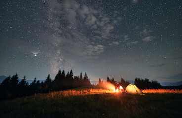 Fantastic view of night starry sky over grassy hill with illuminated camp tents and couple near campfire. Hikers standing under starry sky. Concept of travelling, night camping and relationship.