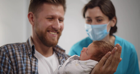 Nurse in protective mask teaching young father to hold newborn son