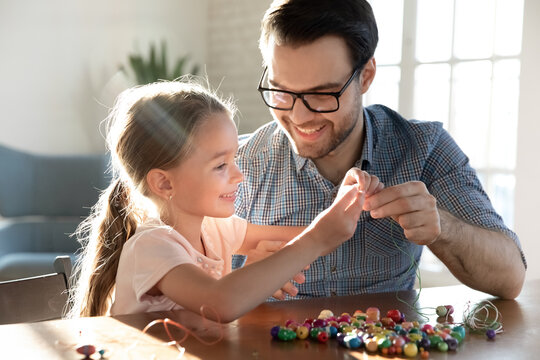 Playful young Caucasian father and little happy daughter have fun making bracelets accessories at home together. Loving dad and small girl child engaged in funny hobby creative activity string beads.
