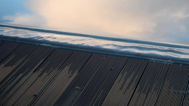 Close up of gentle overflow run off at reservoir. Water is running down under metal grate. Clouds reflected in the water. Accompanying audio.
