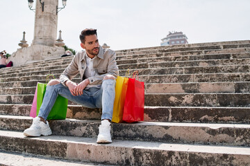 elegant young man with many shopping bags of various colors sitting on a staircase resting after a day of shopping