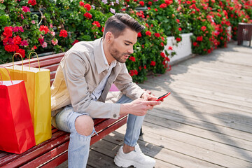 elegant young man sitting on a park bench with many flowers looking at his phone and with many colorful shopping bags next to him, shopping day.