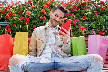 elegant young man sitting on a park bench with many flowers smiling looking at his phone and with many colorful shopping bags next to him, shopping day.
