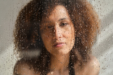 Sensual portrait of young woman taking a shower. Defocused female looks through the glass of the shower stall. Art portrait. Feminine beauty. Skincare wellness.