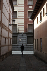 Fototapeta na wymiar Man walking down a narrow, shady street surrounded by walls leading to a brighter one