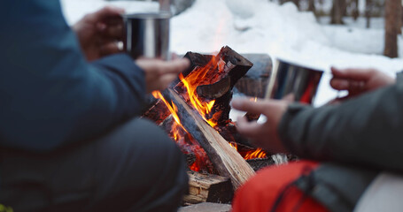 Close up of travelers resting by campfire and drinking hot tea in winter forest