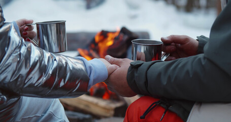 Traveling couple sitting near camp fire with cup of hot coffee and holding hands in winter time.
