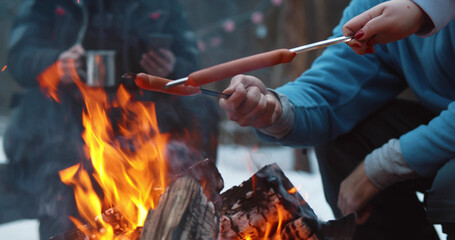 Cropped shot of travelers baking delicious sausages on campfire in winter forest