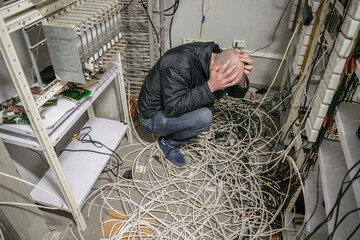 A sad technician sits near a pile of wires and holds his head with his hands in an empty...