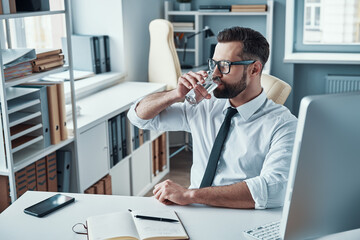 Good looking young man in shirt and tie drinking water while sitting in the office