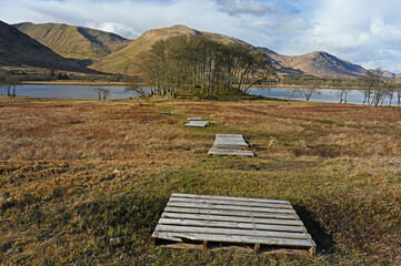 Fototapeta premium View towards Loch Awe in Argyll and Bute, Scottish Highlands with boardwalks forming leading line towards small hilly woodland, loch water and mountains beyond with blue sky and clouds.