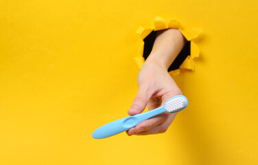 Female hand holds toothbrush through torn hole yellow paper. Oral cavity care. Concept art. Minimalism