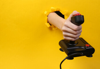 Female hand holds retro joystick through torn hole yellow paper. Concept art.
