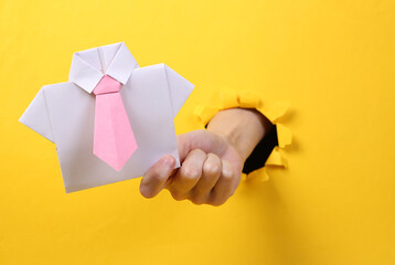 Female hand holds origami shirt with tie through torn hole yellow paper. Concept art. Minimalism.