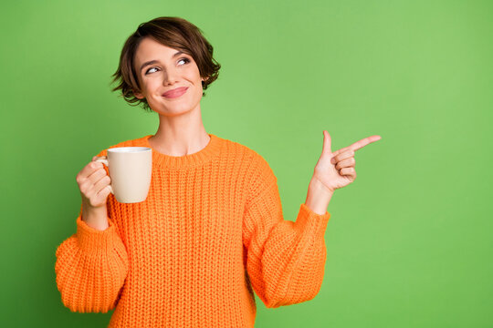 Photo of happy trendy young woman look point index finger empty space hold mug coffee isolated on green color background