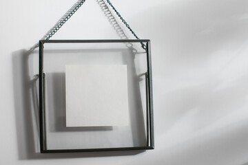 Mockup of black metallic square frame hanging on a chain against a white wall under sunbeams. There are transparent glass installed instead of passepartout