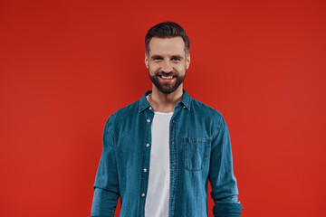 Handsome young man in casual clothing smiling and looking at camera while standing against red background