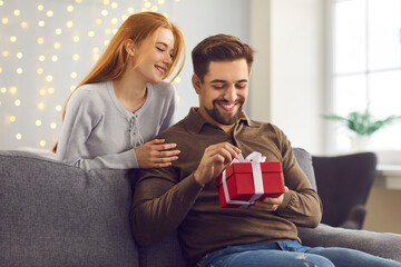 Young happy man boyfriend opening present box from his loving smiling girlfriend during Valentines day celebration at home together. Love, Valentines day, togetherness, dating concept