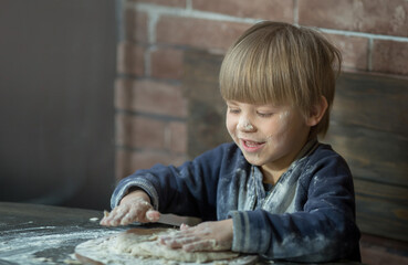 Fototapeta na wymiar a small child boy kneads the dough, he is all soiled in flour, helps his mother to cook. Brick and wood background, copy space