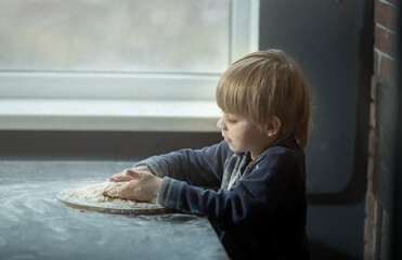 Fototapeta premium a small child boy kneads the dough, he is all soiled in flour, helps his mother to cook. Brick and wood background