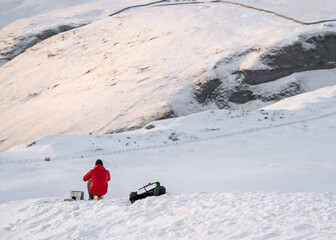 Mountain climber in red coat on white snow covered mountain hill taking break to keep warm eat and drink. Looking at hillside in distance during winter severe weather sat on fold out seat with stove.