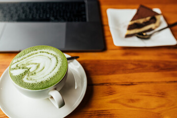 Top view of matcha coffee cup with piece of cake and laptop on brown textured table