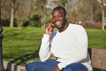 Naklejka na ściany i meble Happy black man talking on smart phone sitting in a park on a sunny day. 