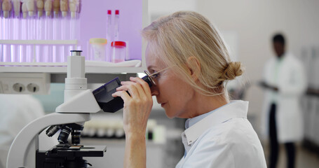 Caucasian mature female scientist looking under microscope analyzing sample.