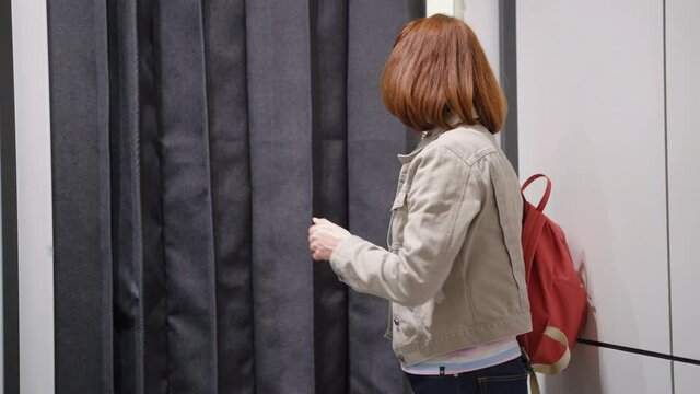 Woman in mask and eyeglasses using smartphone by dressing room, then peeping behind curtain. Female friends shopping together at clothes store and one trying on new outfits. Concept of consumerism