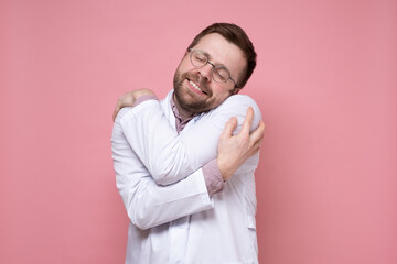 Cute charming doctor is alone, he hugs himself, smiles and dreams with eyes closed. Pink background.