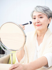 Senior asian woman applying foundation to her cheek with a makeup brush while sitting alone in front of a mirror.