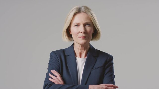 Studio portrait of mature businesswoman with a serious expression against grey background wearing business suit folding her arms and looking at camera - shot in slow motion