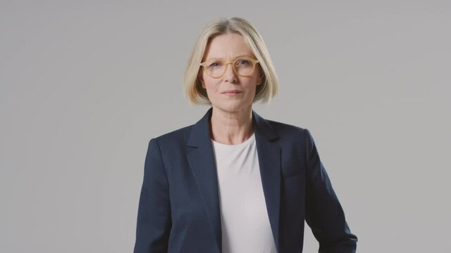 Studio portrait of mature businesswoman wearing designer glasses with a serious expression against grey background in business suit looking at camera - shot in slow motion