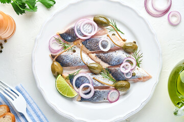 Salted herring with spice, herbs and onion on white plate on light stone background with copy space. Marinated sliced fish. Food with healthy unsaturated fats, flat lay