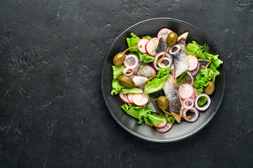 Salted herring with spice, capers, herbs and onion salad on black plate on dark background with copy space. Marinated sliced fish. Food with healthy unsaturated fats, flat lay