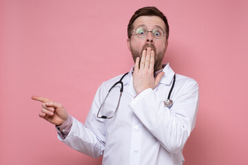 Shocked doctor with a stethoscope around neck, frightened, points finger to the side, looks at the camera and covers mouth with hand.