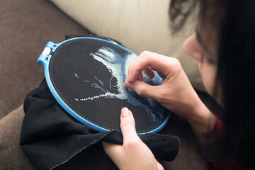 A woman embroiders a picture with threads in cross while lying on the sofa.