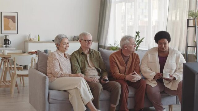 Medium long of four diverse senior friends sitting on couch in living room, watching TV, chatting laughing