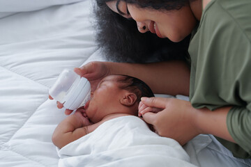 Portrait of African newborn baby boy wrapped in blanket drinking milk in plastic bottle sleeping on white bed. Young big mother feeding bottle of milk to cute infant while holding his hand at bedroom