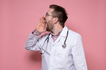 Young doctor with a stethoscope around neck is calling someone, he screams loudly with hand to mouth and looks away. Pink background.