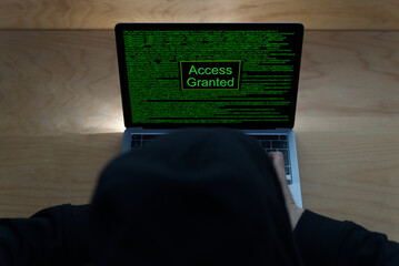 Person with black hoodie in front of laptop computer. Black screen with green signs and text access granted. Photo taken April 12th, 2021, Zurich, Switzerland.