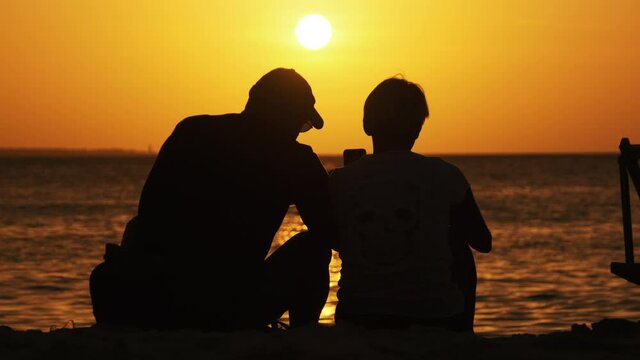 Silhouette Couple of middle-aged man and woman at sunset sitting on the beach by the Ocean, Zanzibar. Rear view of people looking at the sundown over the water on paradise resort. Slow Motion. 4K.