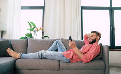 Young handsome happy bearded man in glasses is using smartphone for surfing in net, typing, and chatting, playing games, or working with some apps while relaxing on the couch at home