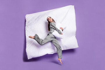 Full length body size photo of woman jumping on bed embracing pillow in grey sleepwear isolated pastel violet color background