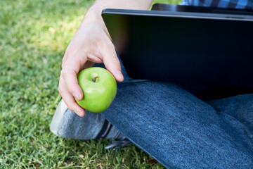 Unrecognizable young man holding an apple while using his laptop outdoors. Conceptos of healthy eating and online working or studying., digital nomadism.