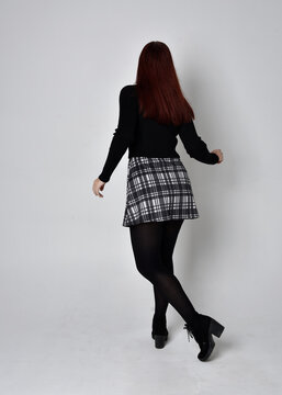 Full length portrait of a red haired  girl wearing black turtleneck sweater,  plaid skirt and boots.  Standing pose with back to the camera against a studio background.
