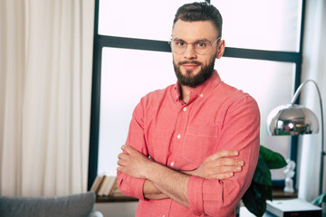 Naklejka na ściany i meble Handsome confident and happy bearded man in eyeglasses and shirt is standing with crossed arms and looking on the camera