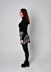 Full length portrait of a red haired  girl wearing black turtleneck sweater,  plaid skirt and boots.  Standing pose in side profile against a studio background.